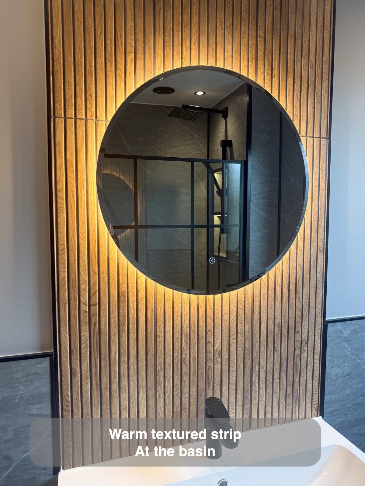 Vertical wood-look ribbed tile strip behind circular backlit mirror and white basin.