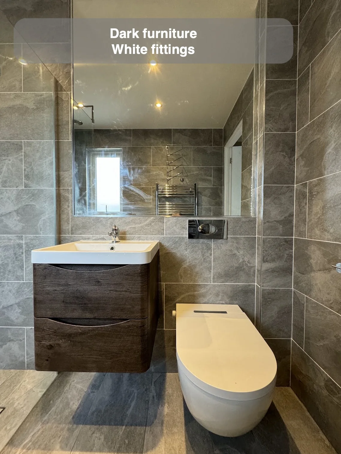 Dark curved drawer fronts beneath rectangular white basin beside wall-hung toilet and grey tiles.