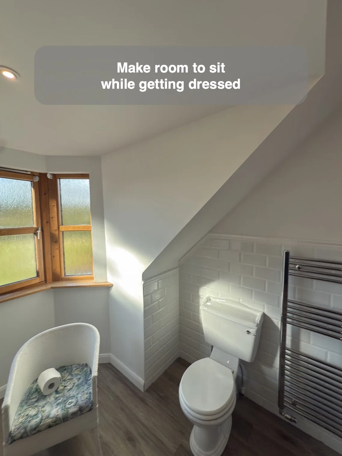 Compact dressing chair beside a window beneath a sloping bathroom ceiling