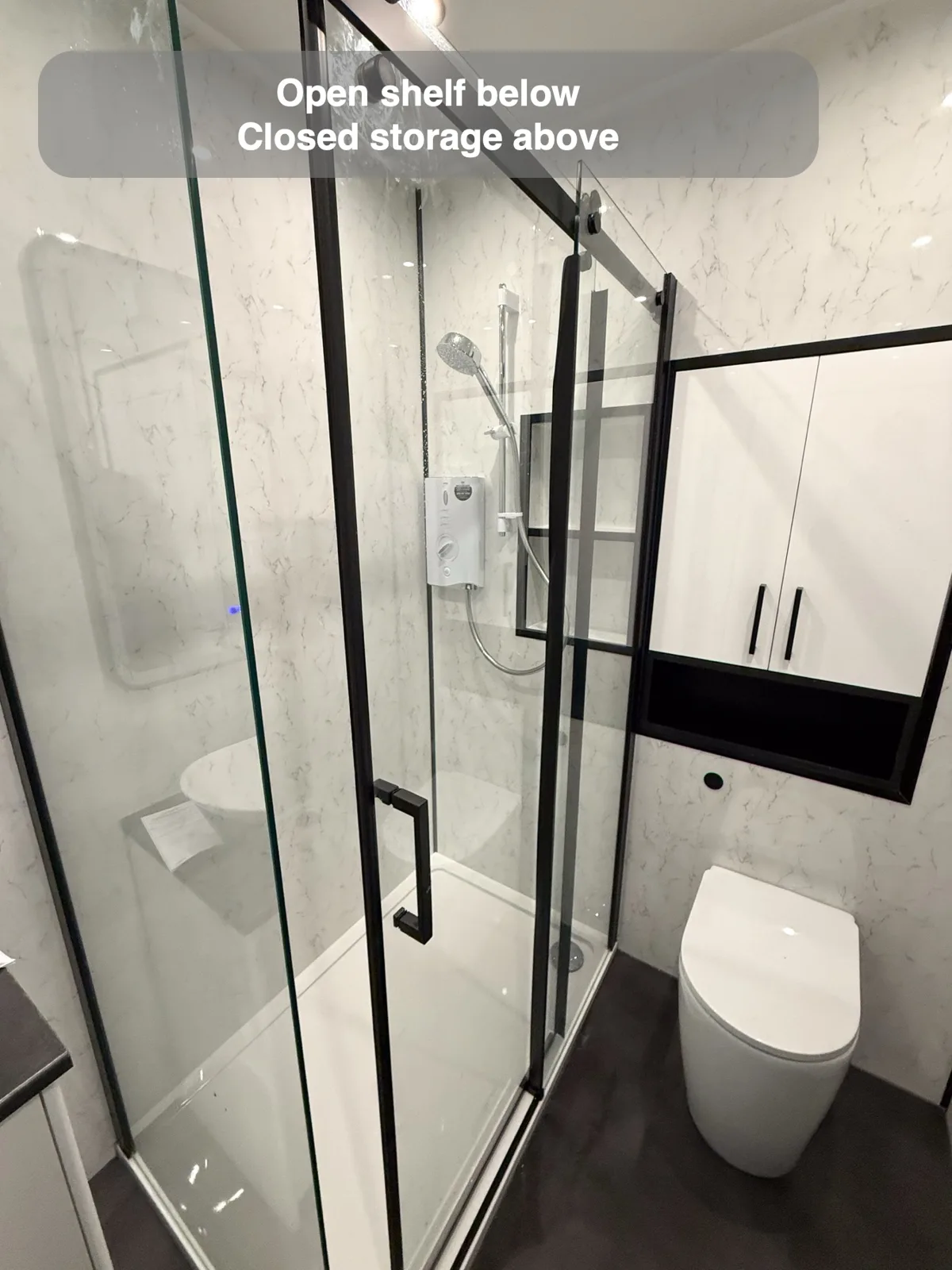 White cupboard above toilet with dark-framed open shelf underneath and black shower frame.