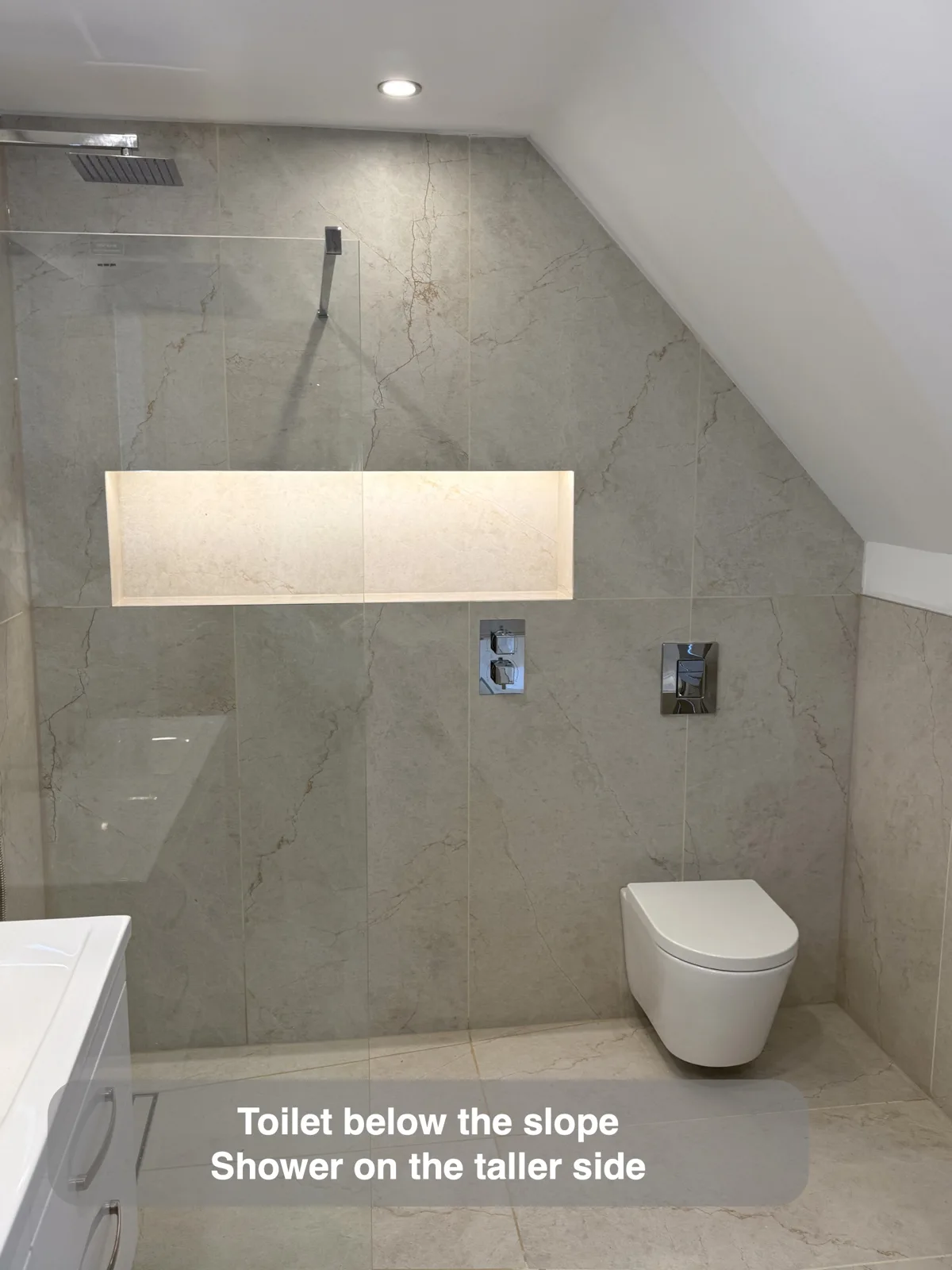 Wall-hung toilet beneath roof slope; shower at taller side of same tiled wall.