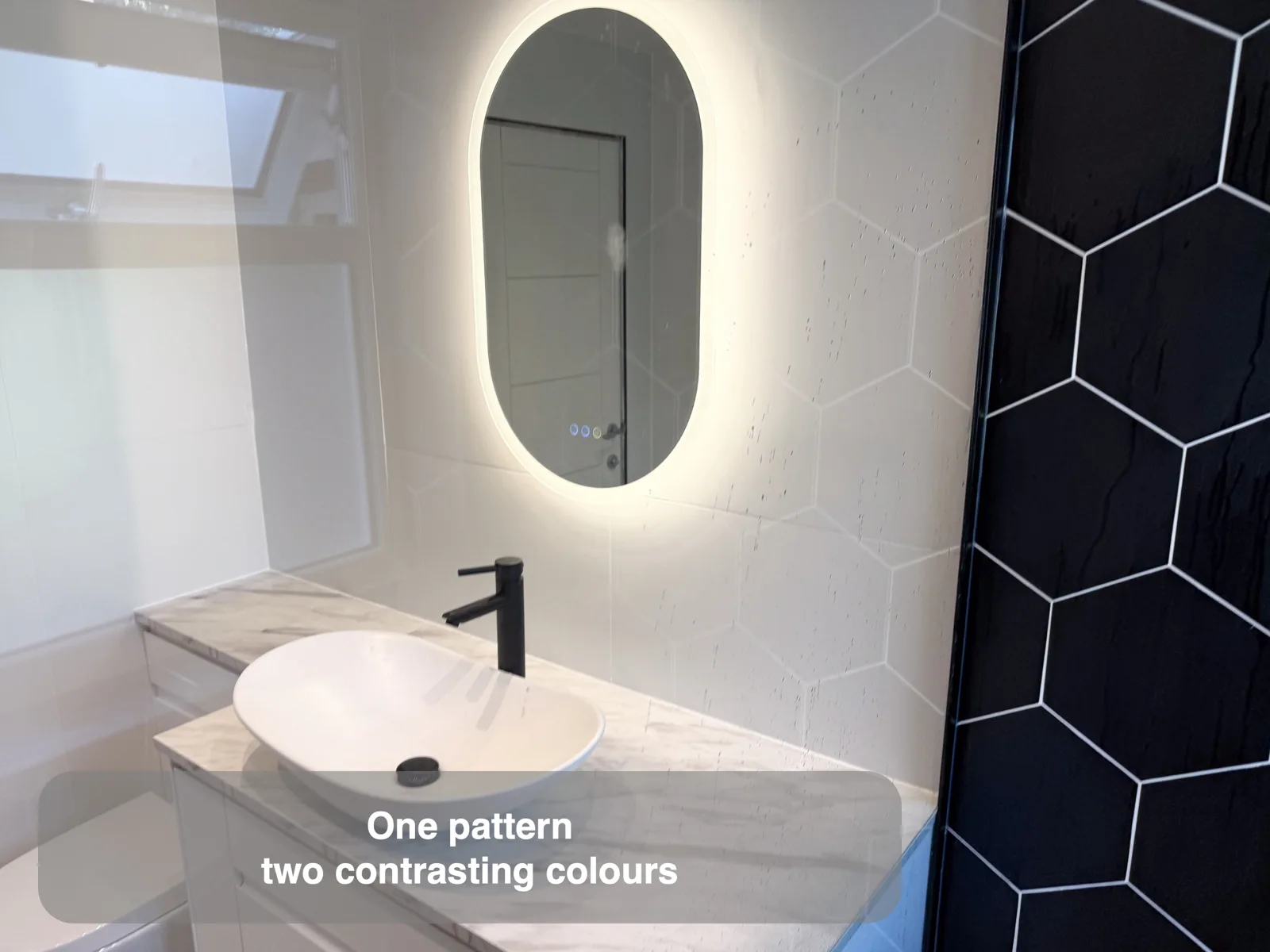 Pale hexagonal basin wall beside a black hexagonal shower wall, with an oval lit mirror and white vessel basin.