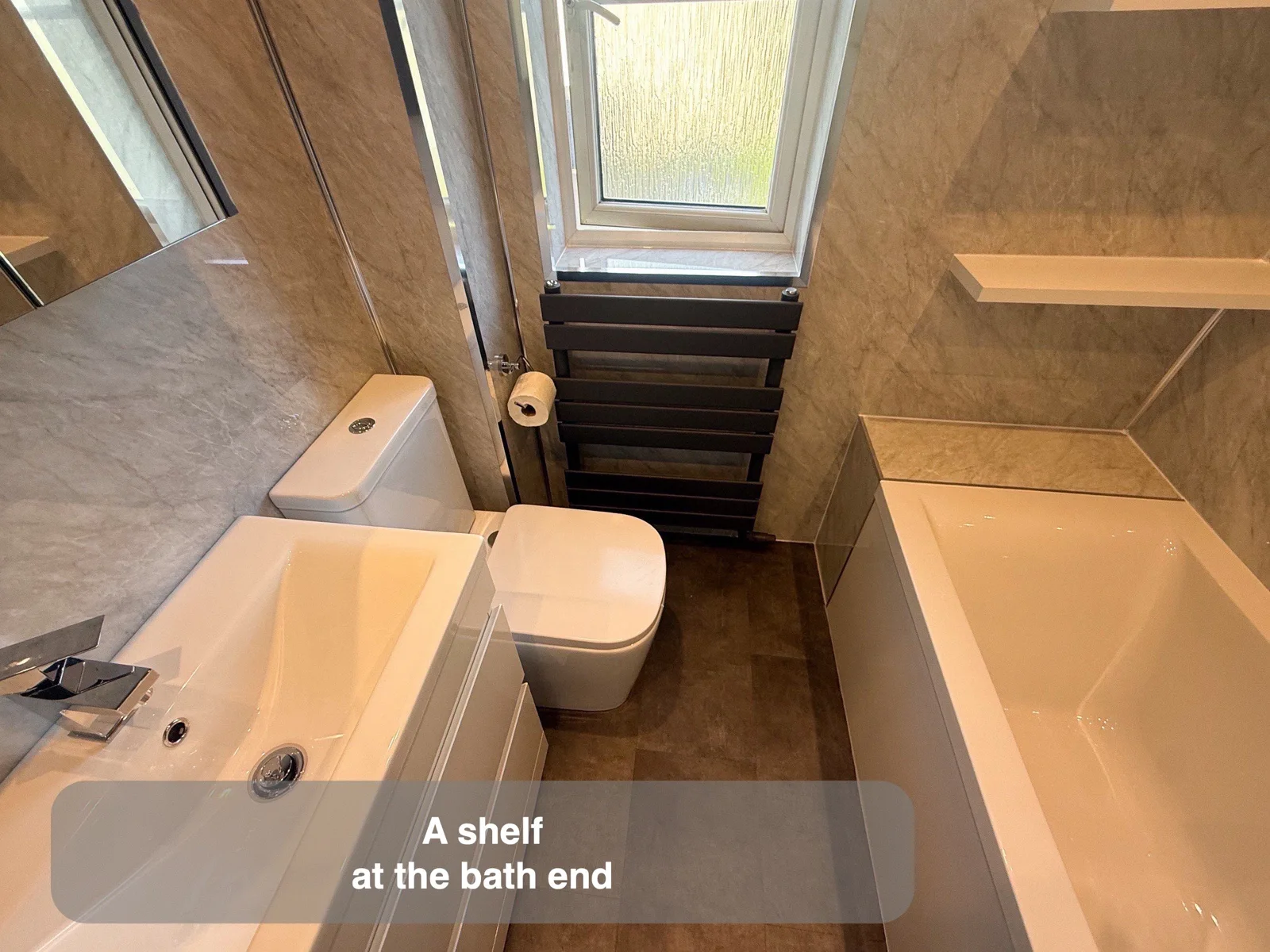 White bath ending beneath a pale narrow wall shelf and a deeper ledge, opposite basin furniture and beside a window.