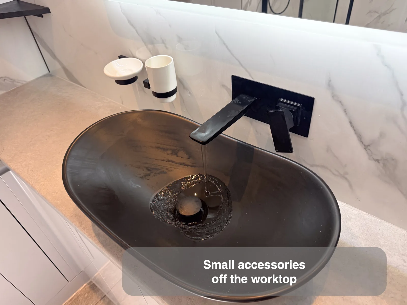 White soap dish and tumbler in black wall brackets beside a wall tap over a dark vessel basin.