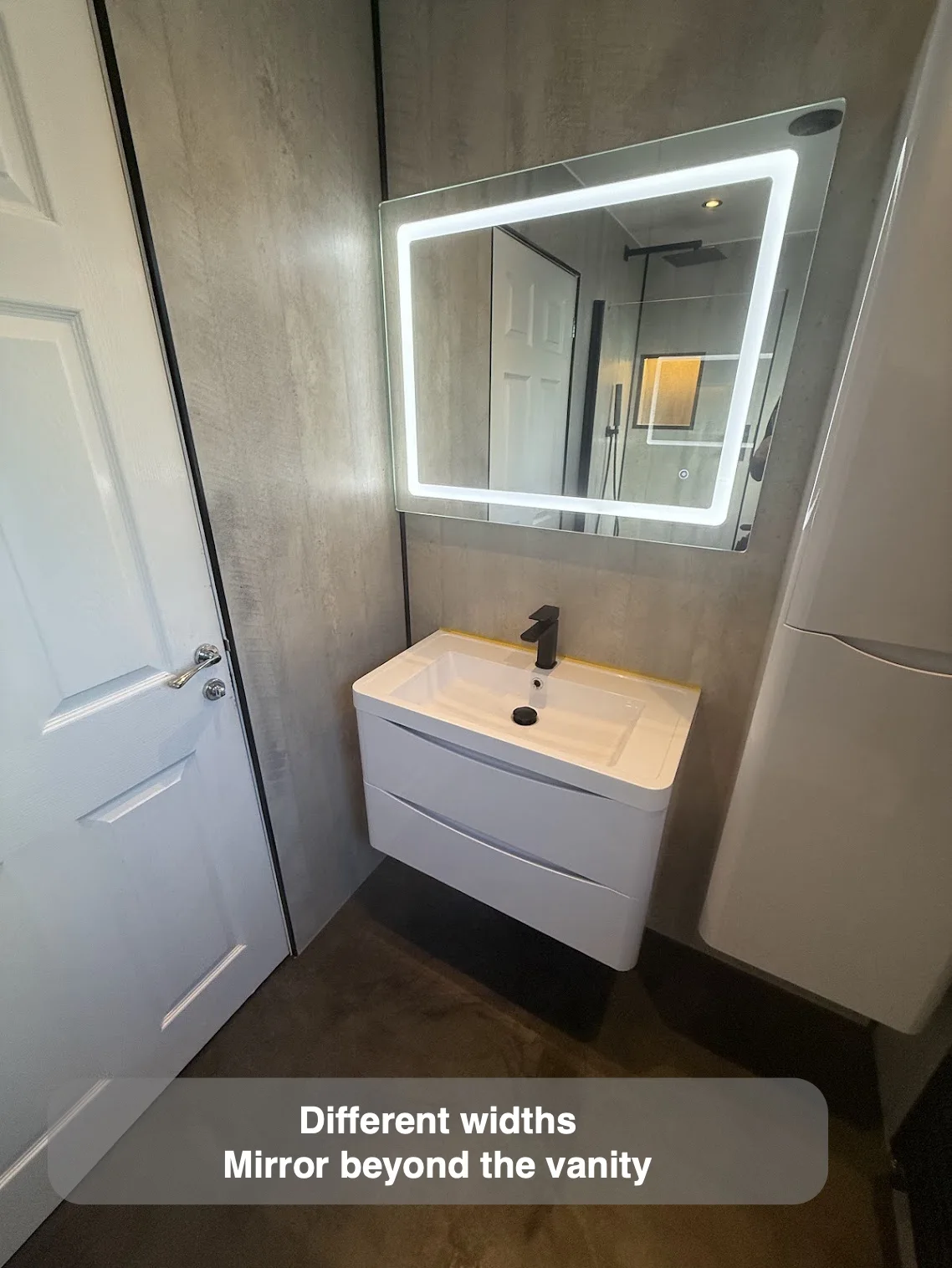 Illuminated wide mirror above a compact white basin unit beside a door.