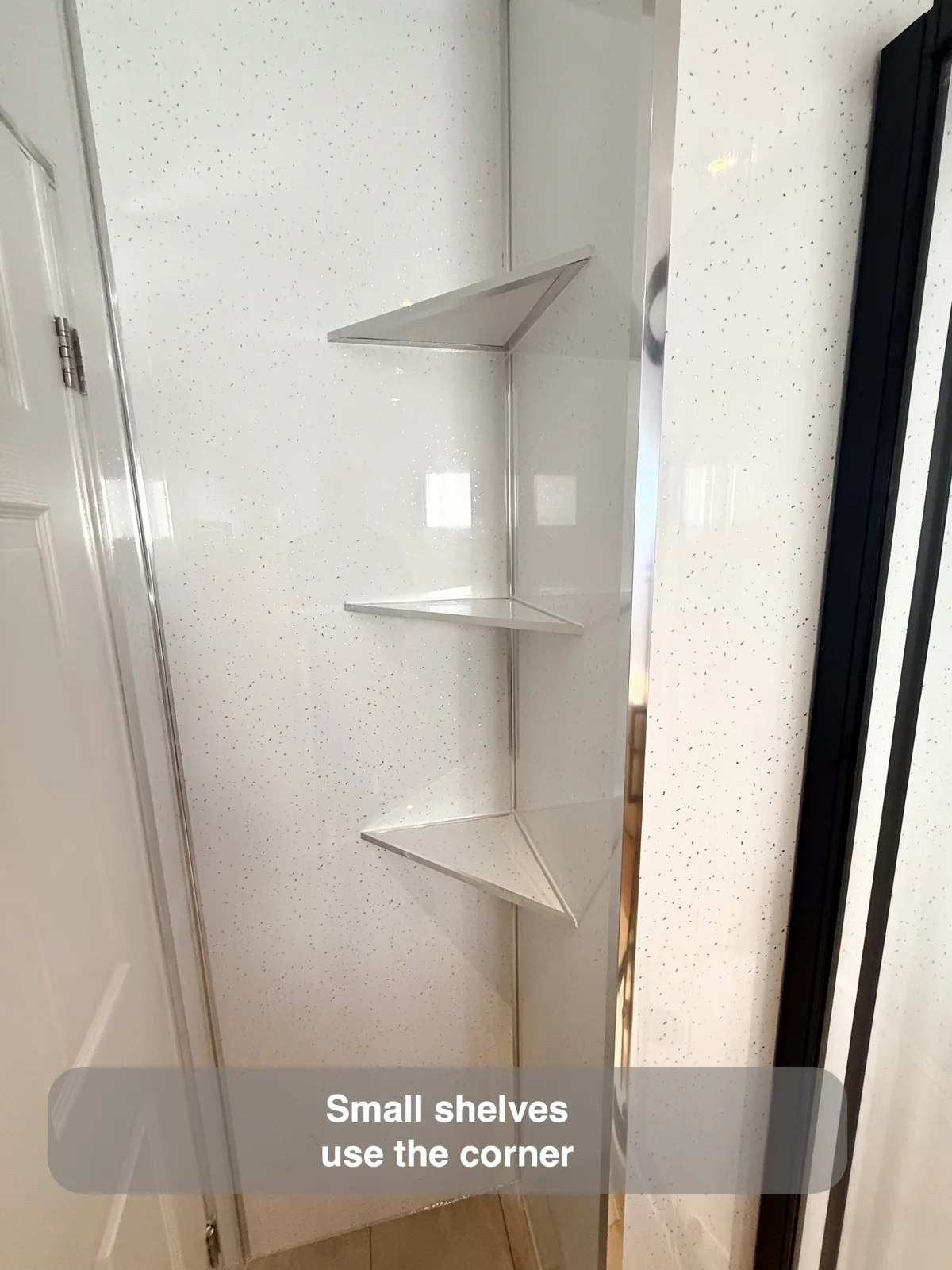 Three triangular pale shelves in a narrow wall corner between a white door and black shower frame.
