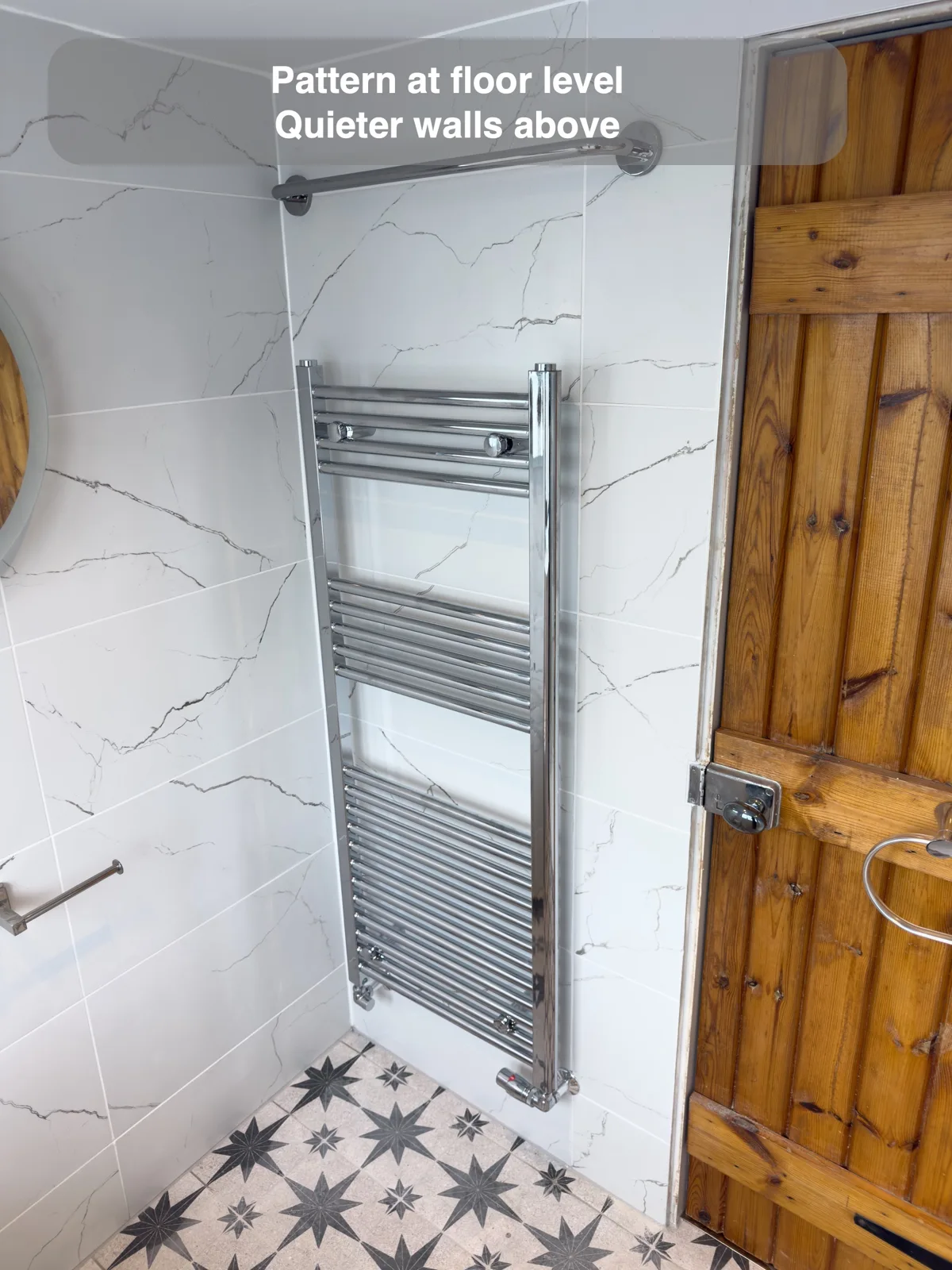 Patterned floor below pale veined walls, beside a chrome towel rail and timber door.