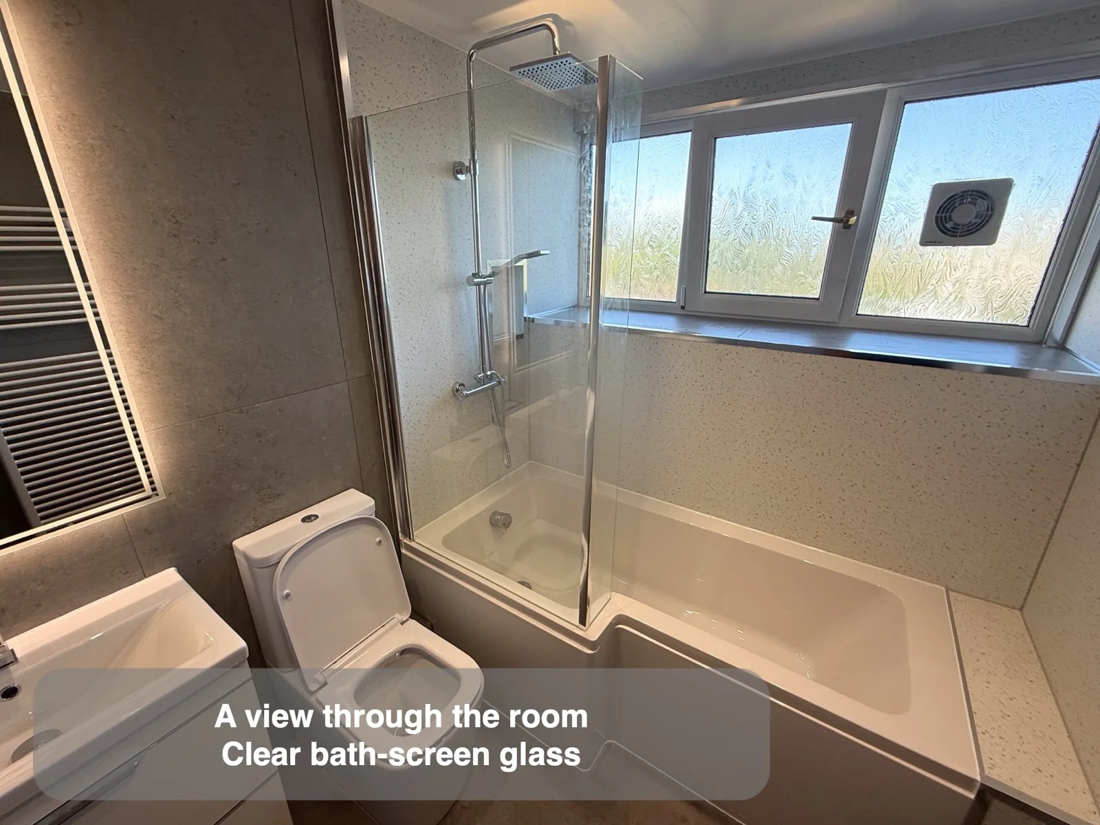 Bath, clear shower screen and window in a pale bathroom.