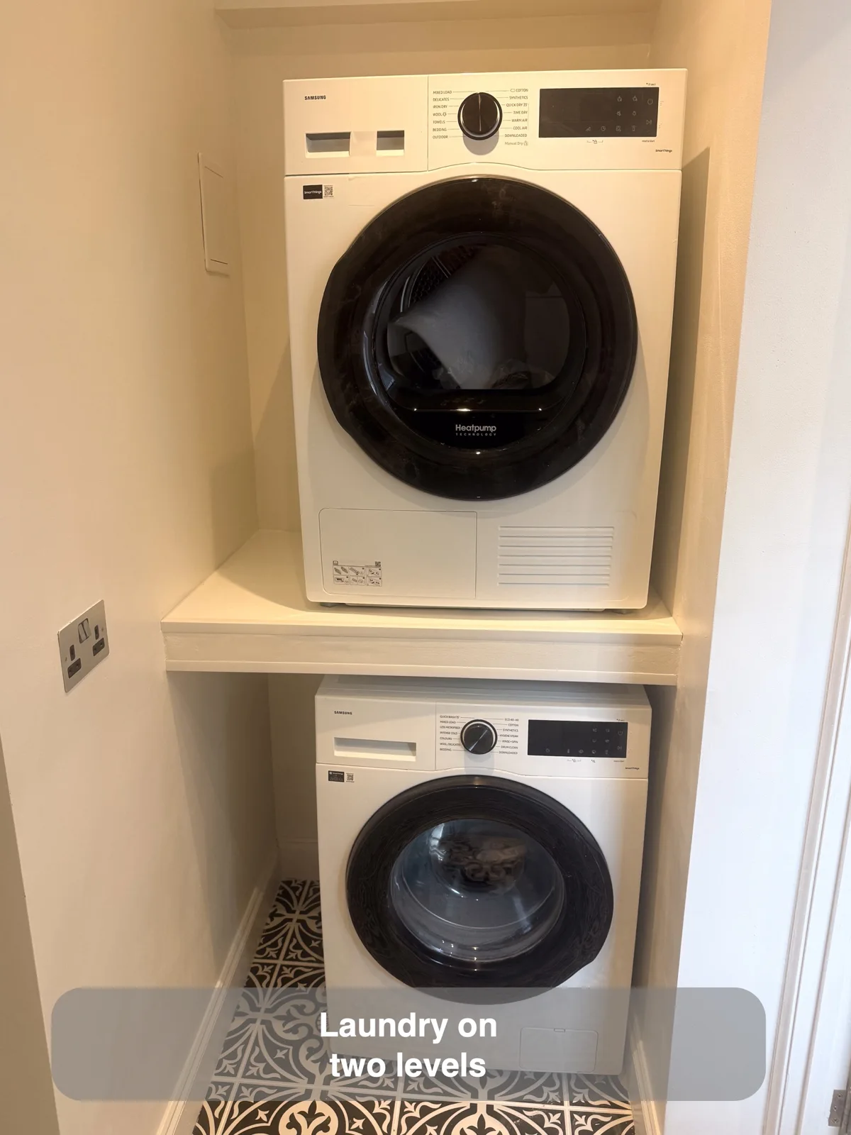 White dryer on a broad shelf above a white washing machine in a narrow alcove with patterned flooring.