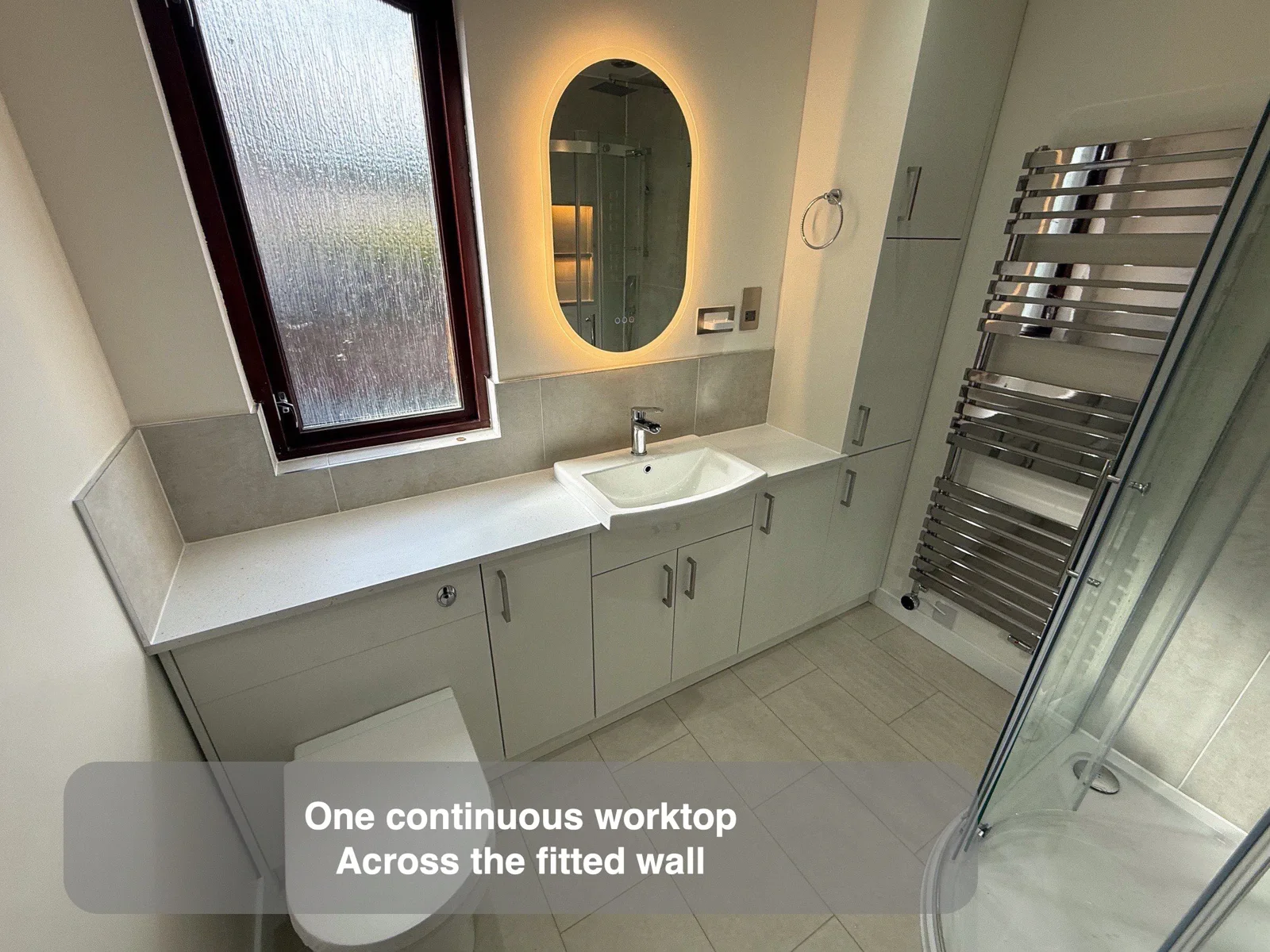 Continuous white worktop below tall window and above toilet/cupboards to basin.